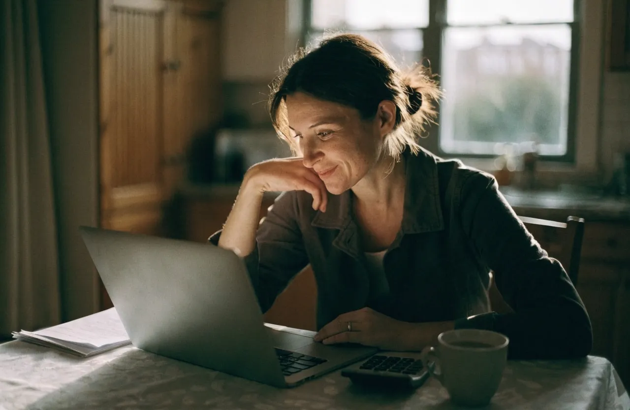 A woman at her kitchen table working out course costs on a laptop and calculator.